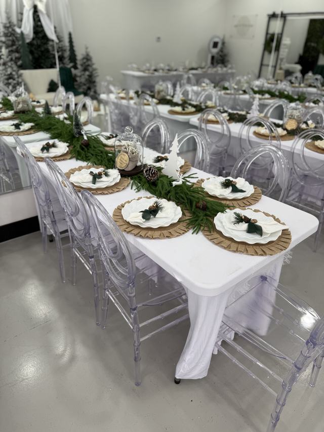 Long tables dressed in white linens with greenery runners, woven chargers, and clear chiavari ghost chairs