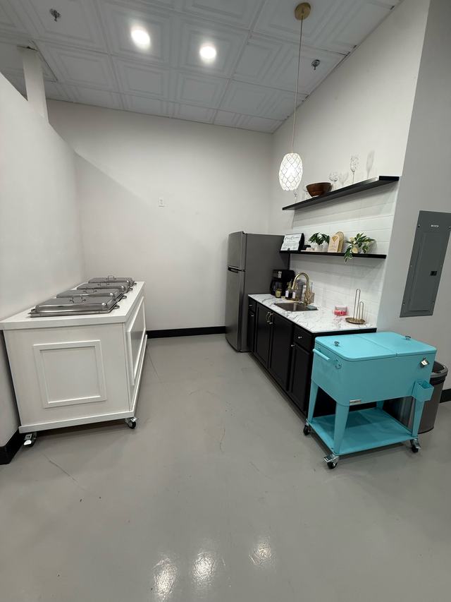 Kitchenette prep area with fridge and shelving, a white food cart, and a teal mobile drink cooler