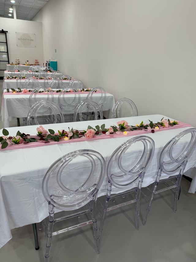 Long tables with white linens, pink runners, rose garlands, and clear ghost chairs